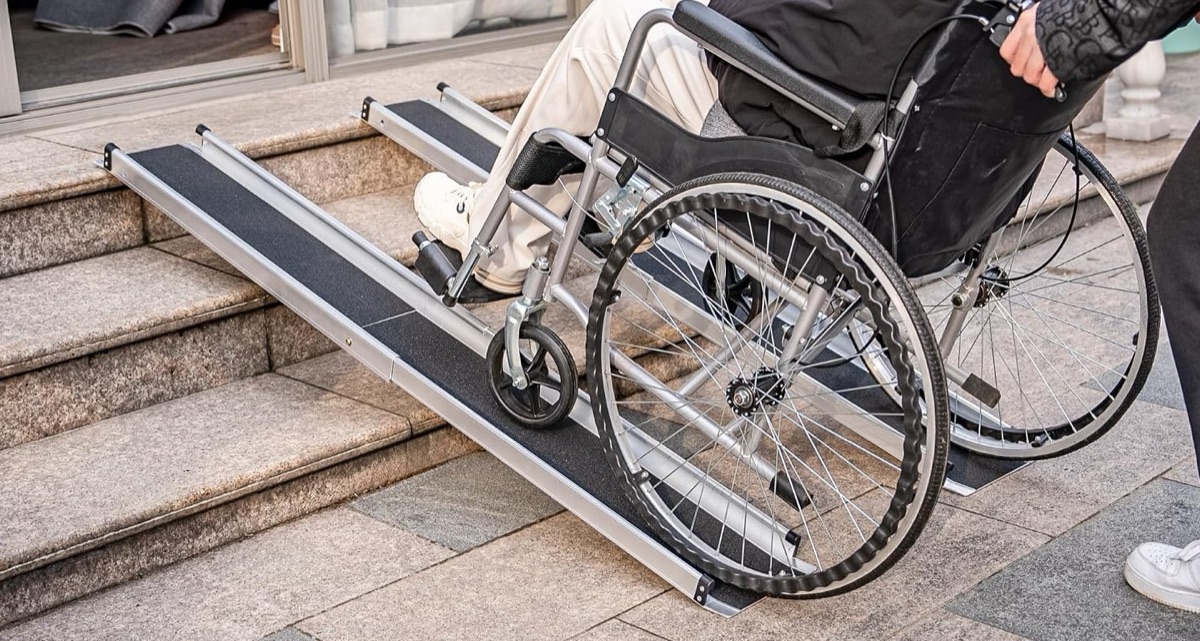 A wheelchair client faces forward going up a portable ramp over stone steps, pushed from behind