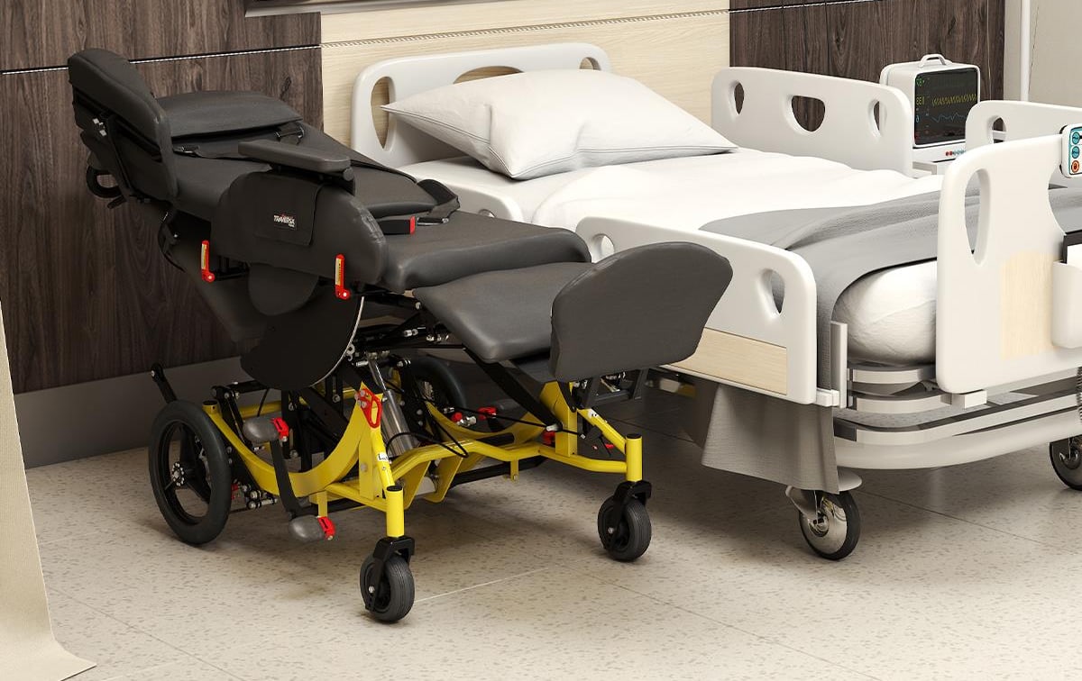 A reclined, height-adjustable transport chair positioned beside a hospital bed, ready for a side-to-side transfer